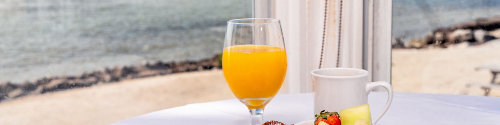 A table with a plate of pastries, a bowl of fruit, a glass of orange juice, and cutlery, set by the beach.