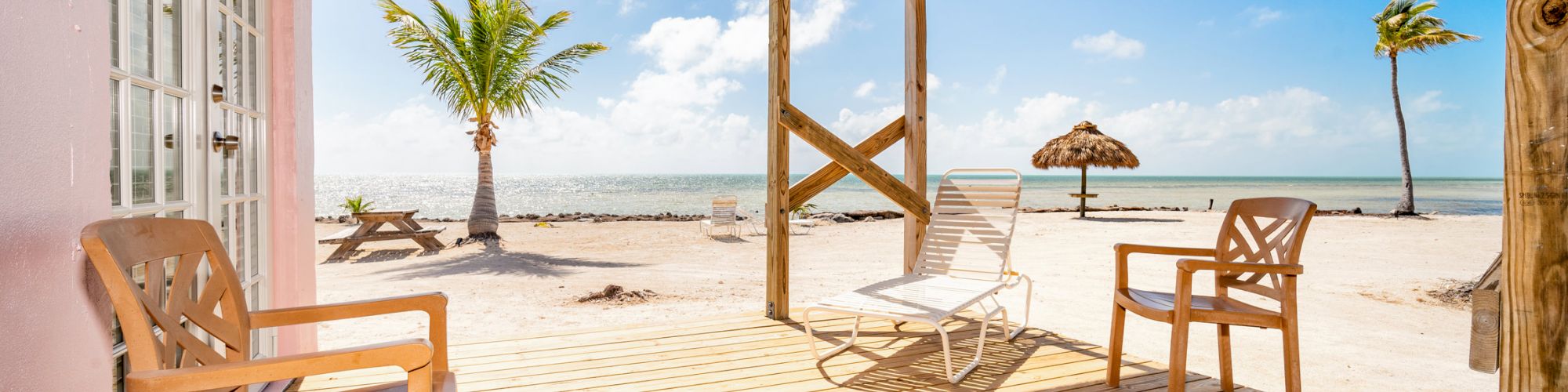 A beachfront deck with wooden chairs, a thatch umbrella, and a palm tree, overlooking a sandy beach and clear blue sky ending the sentence.