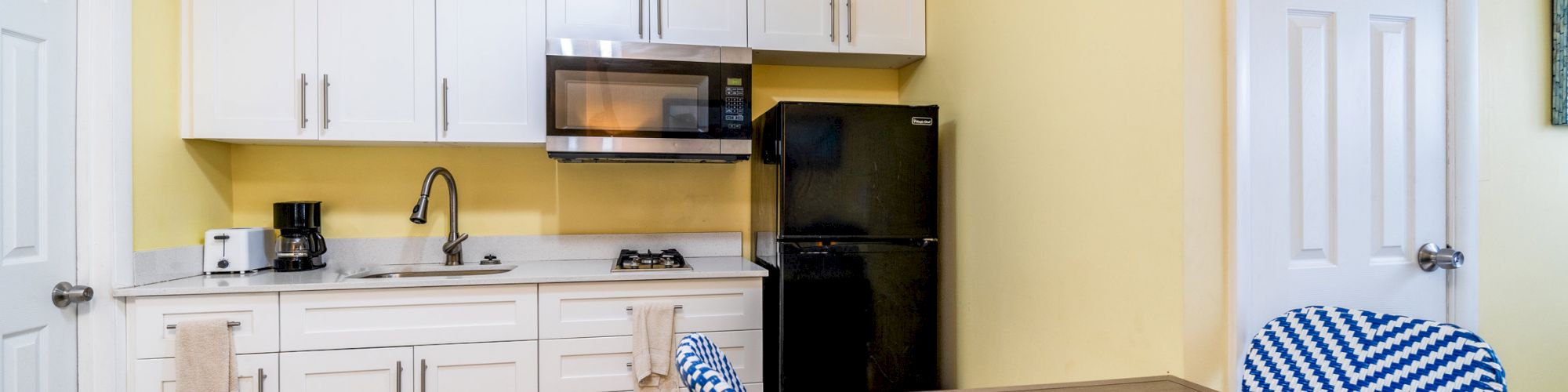 A small kitchen with white cabinets, a black refrigerator, a microwave, a dining table, and blue-striped chairs in a room with yellow walls.