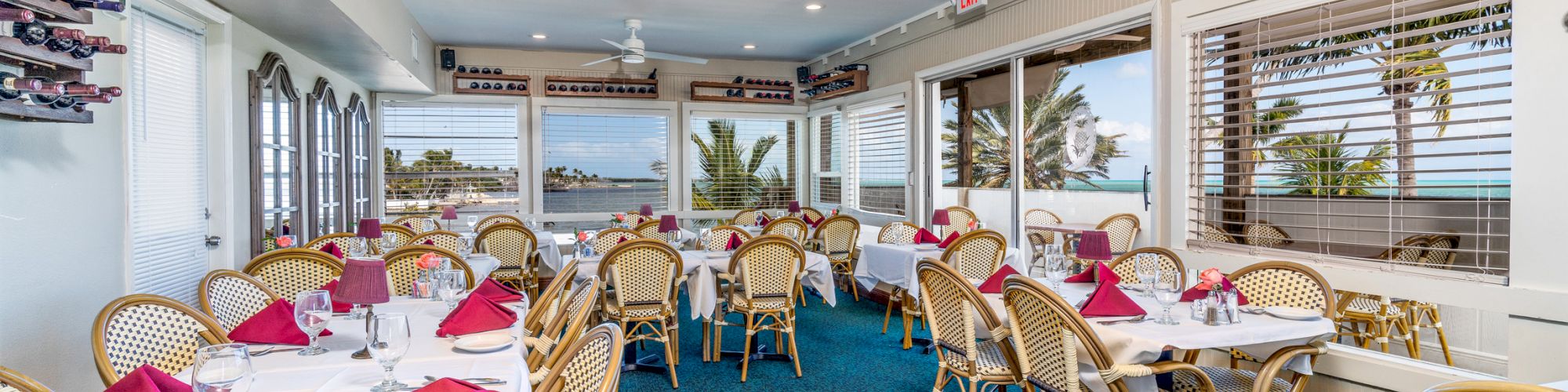 A bright restaurant with wicker chairs, white tablecloths, and red napkins, featuring large windows offering a scenic view outside, can be seen.