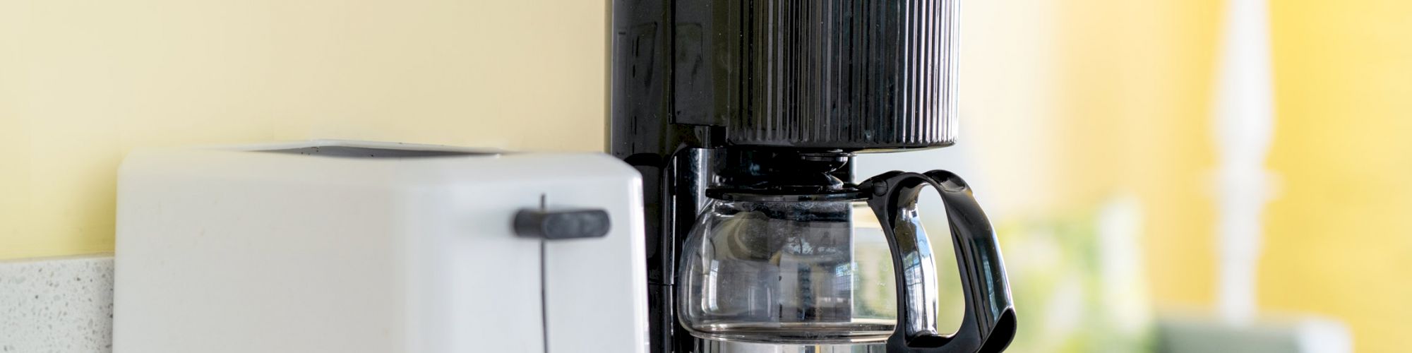 A white toaster and a black coffee maker are placed on a kitchen counter with a lamp and some furniture visible in the background.