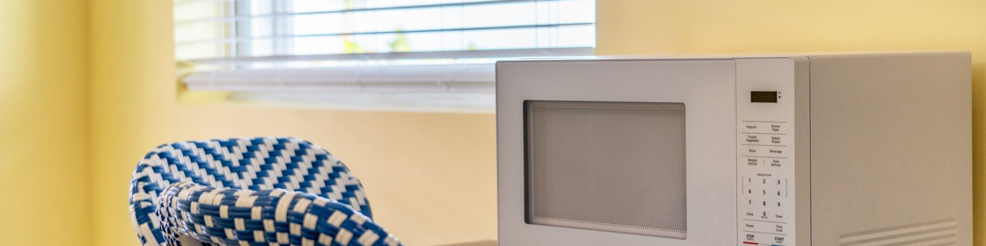 A microwave sits on a wooden table next to a window with blinds and a blue and white patterned chair in a room with yellow walls.