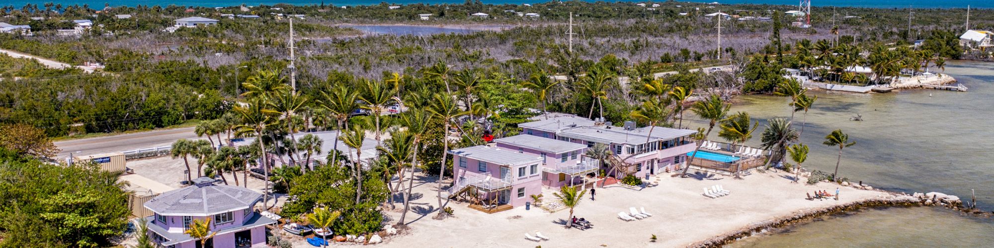 An aerial view of a beachfront property with buildings, palm trees, and a pathway adjacent to the ocean, under a blue sky and scattered clouds.