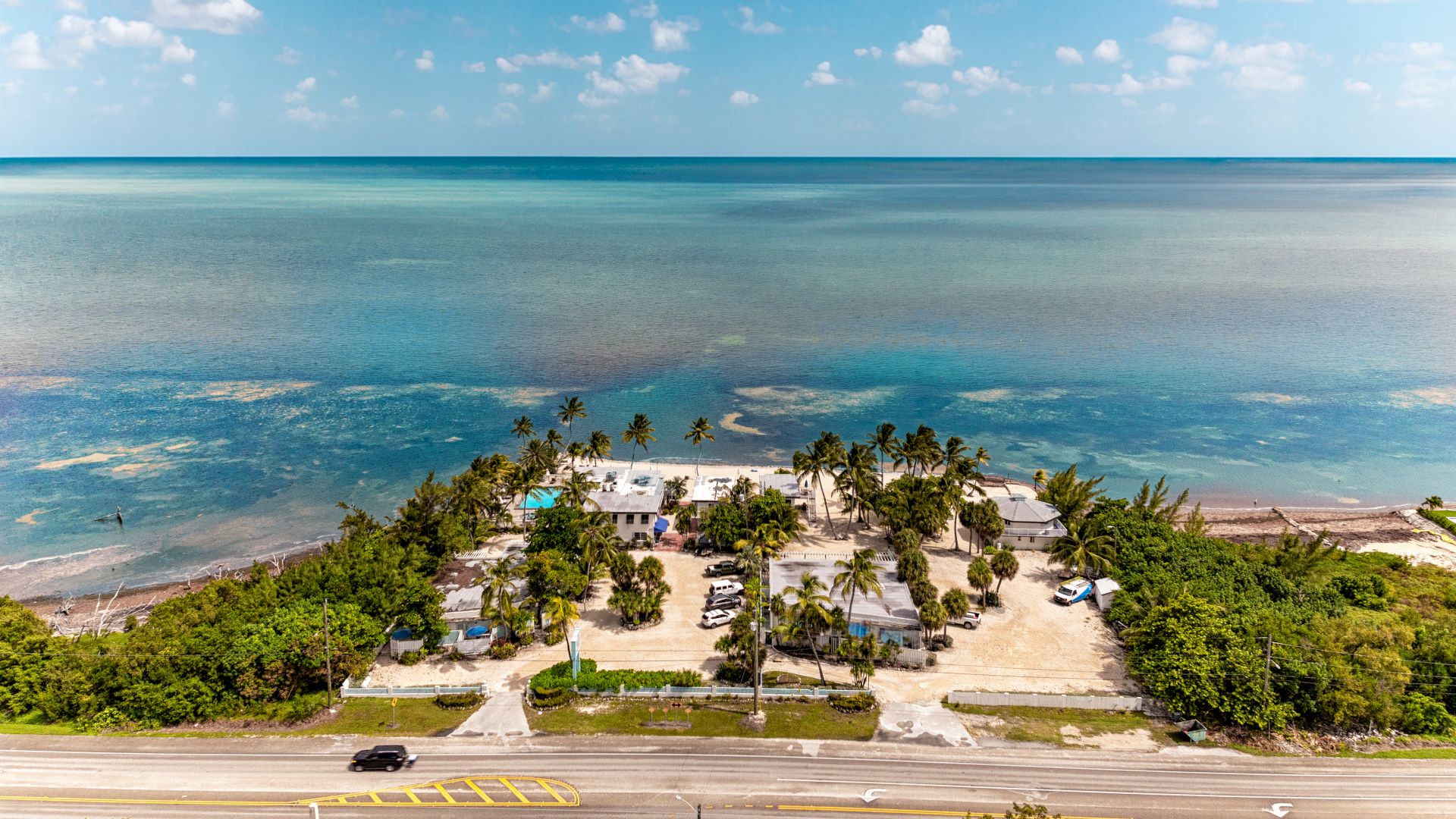 A coastal scene with a road, buildings, trees, and a vast ocean under a partly cloudy sky, creating a serene and tropical atmosphere.