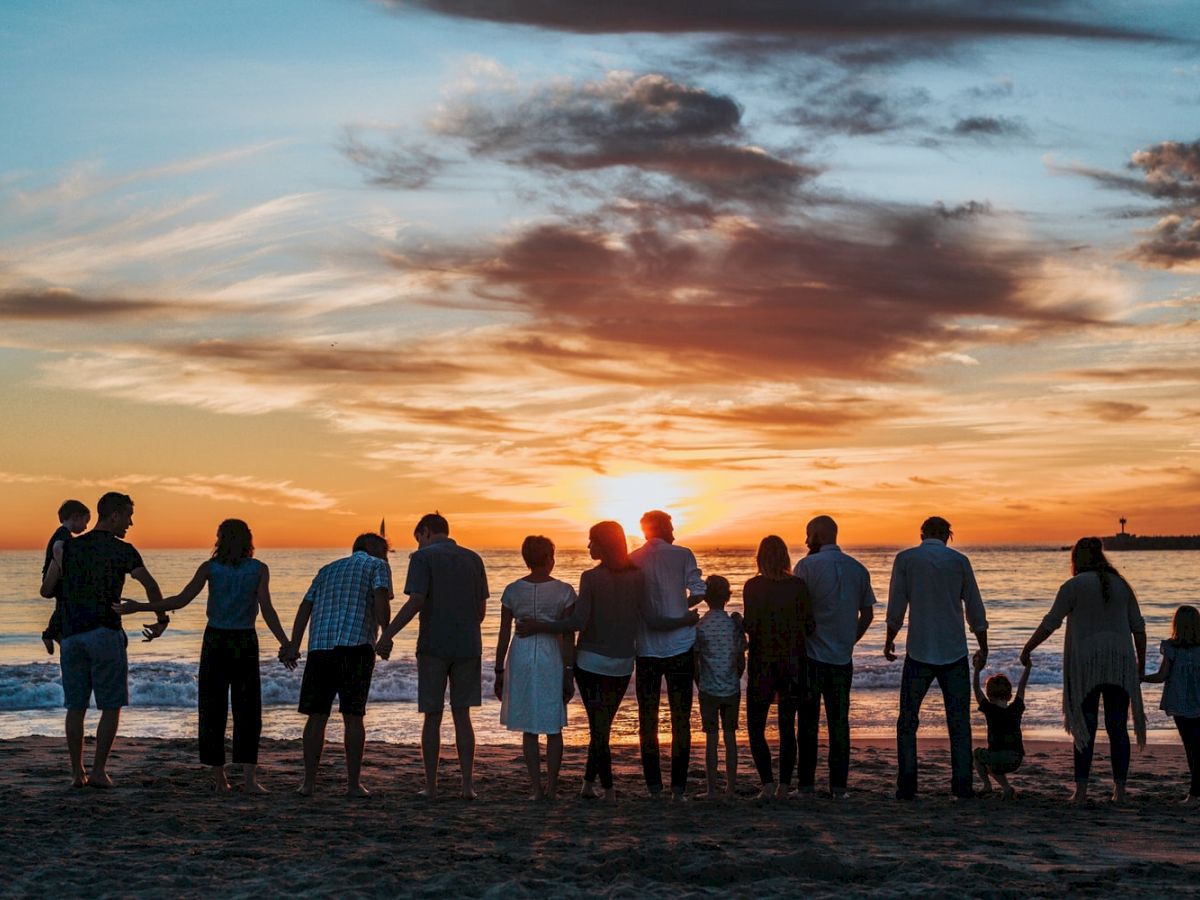 A group of people holding hands and standing on a beach, facing a beautiful sunset and showcasing a sense of togetherness and harmony.