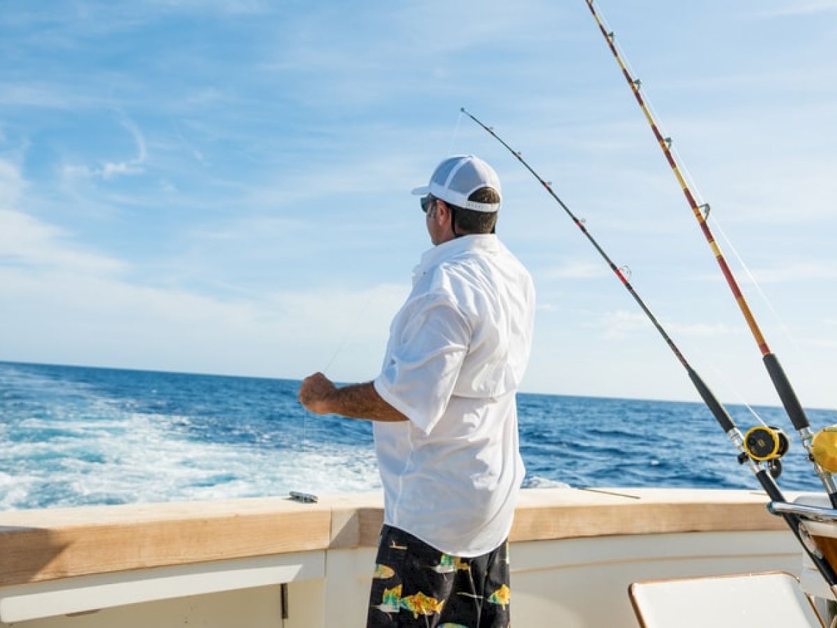 A man in a white shirt and cap stands on a boat holding a fishing rod, with the ocean and two fishing poles in the background.