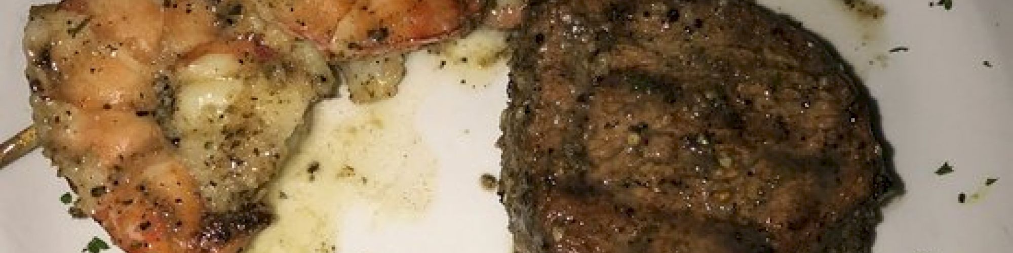 The image shows a plate with grilled shrimp skewers and a seasoned steak filet garnished with chopped herbs.