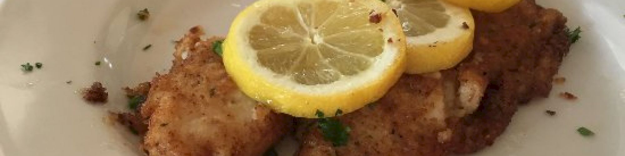 The image shows a plate with fried fish garnished with lemon slices and parsley, a glass of orange drink, and a glass of water on a table.