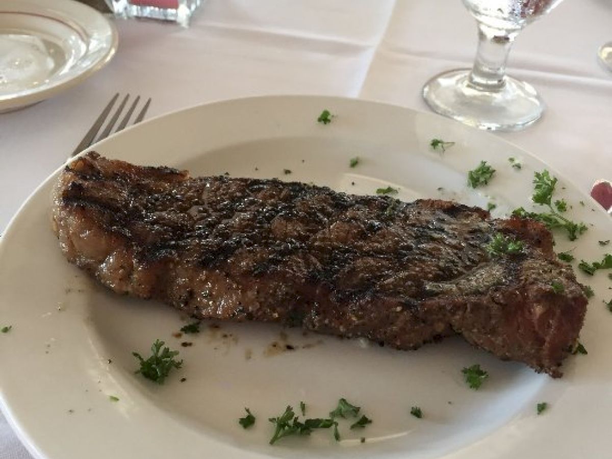 A grilled steak garnished with herbs on a white plate, accompanied by water glasses and other dining essentials on a white tablecloth.