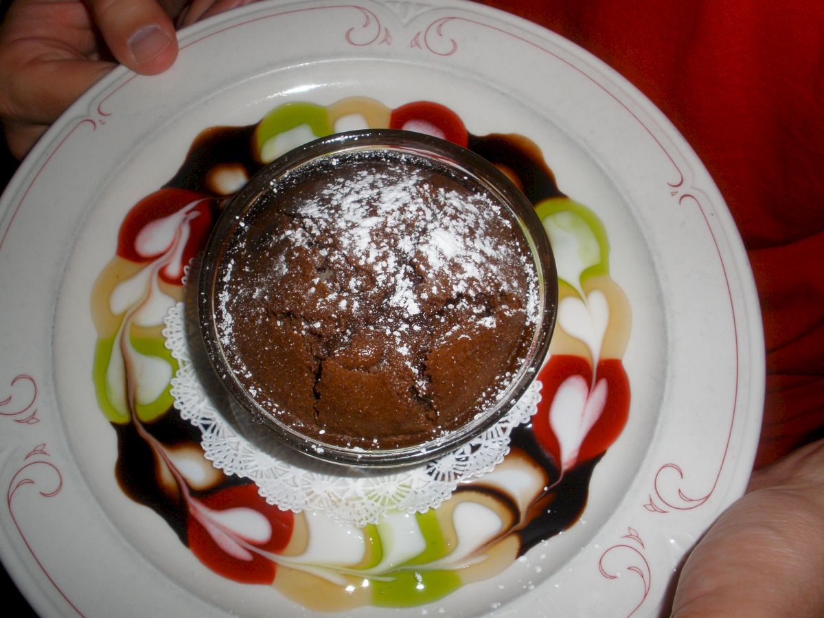 A person is holding a plate with a chocolate dessert topped with powdered sugar. The plate features colorful decorative designs.