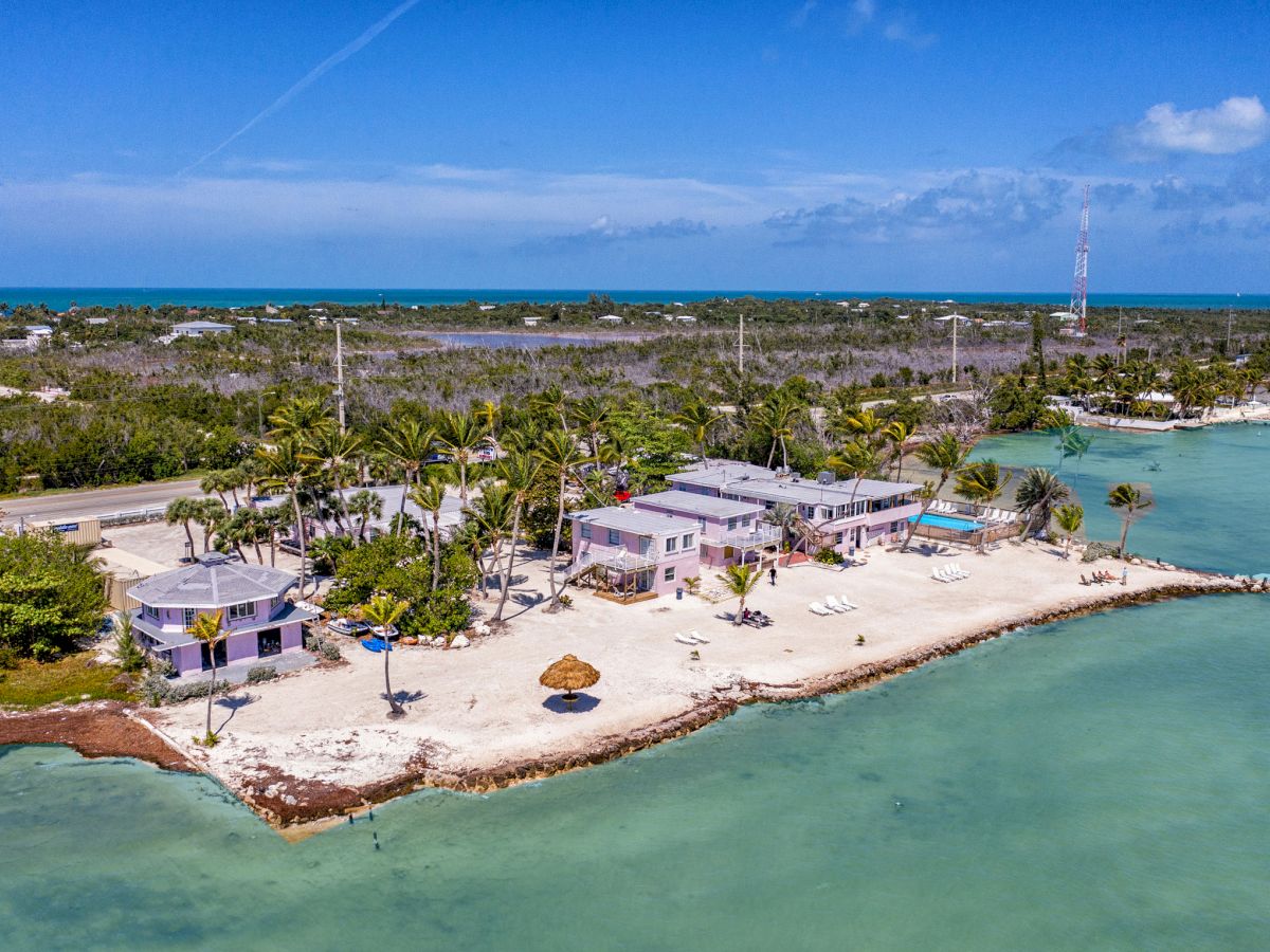 Aerial view of a coastal resort with buildings, palm trees, and a sandy beach bordered by turquoise waters, under a clear blue sky.