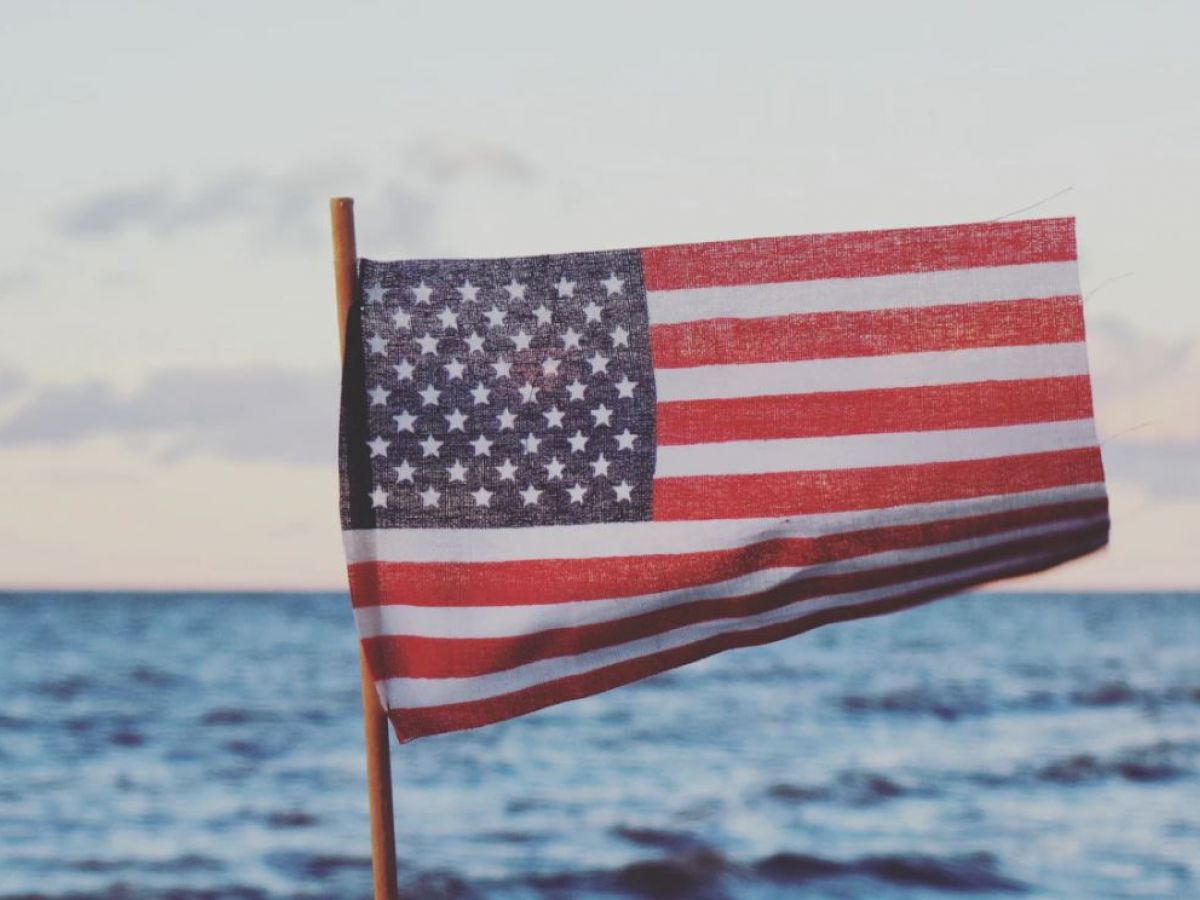 The image shows an American flag on a pole, with a body of water and a partly cloudy sky in the background.