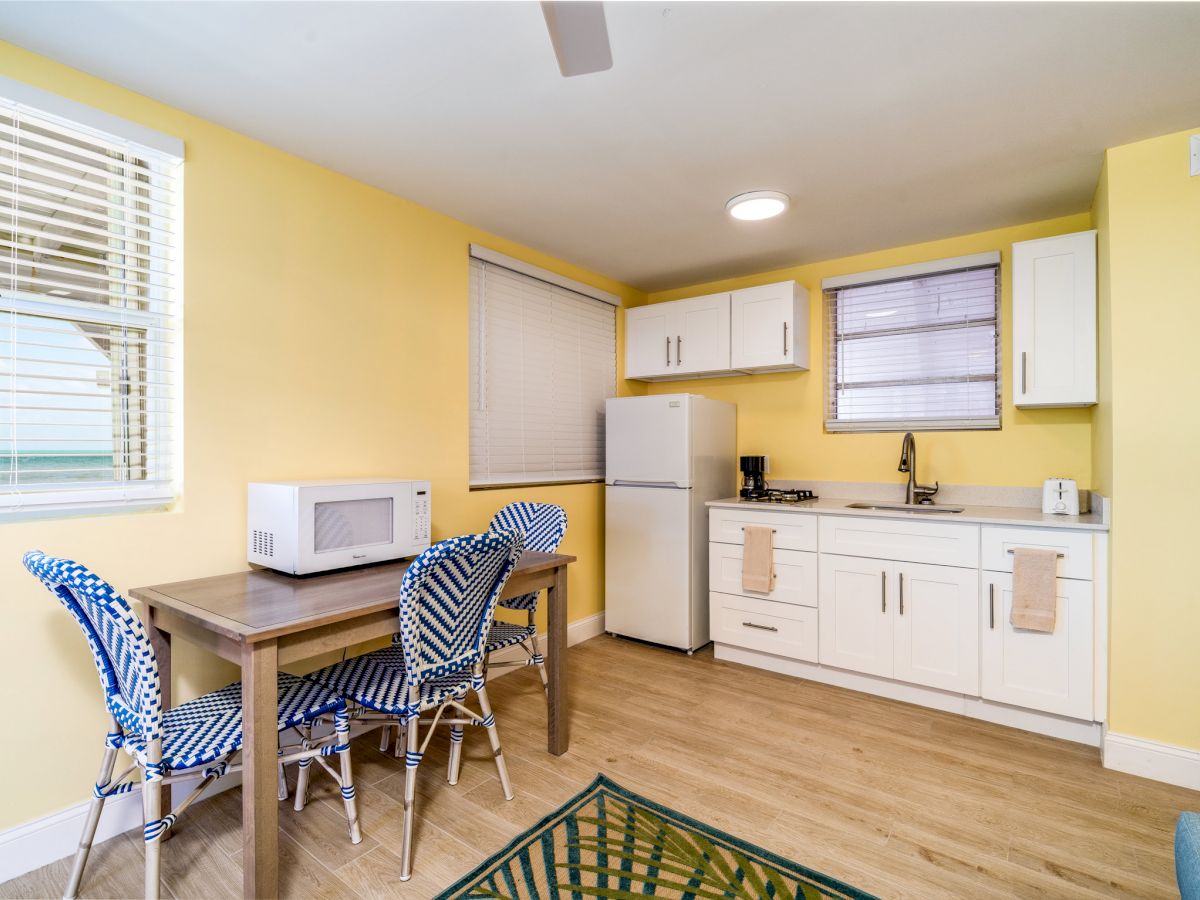 A small kitchen and dining area with white cabinets, a fridge, microwave, dining table, and chairs, in a room with yellow walls and wooden flooring.