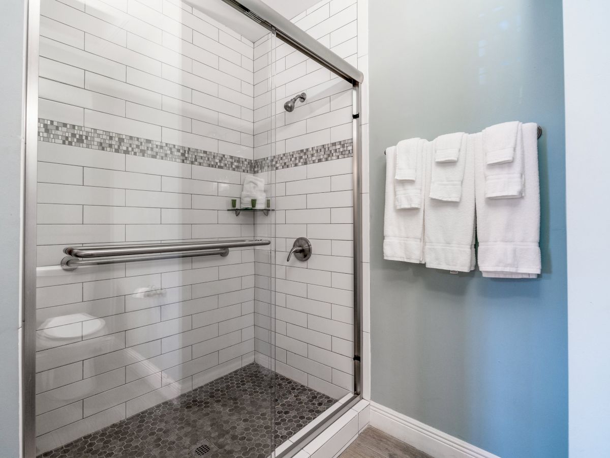 This image shows a modern, glass-enclosed shower with white subway tiles, a horizontal decorative strip, and three white towels hanging on the wall.