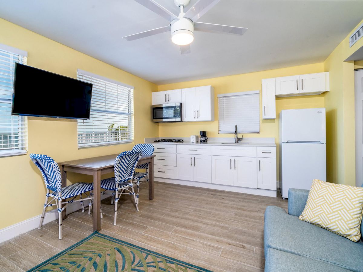 A small, clean kitchen/lounge area with yellow walls, white cabinets, a sofa, dining table with chairs, TV, and a refrigerator.