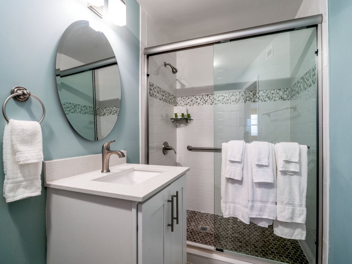 The image shows a modern bathroom with a vanity, mirror, hand towel, and a glass-enclosed shower with white towels hanging on the grab bar.