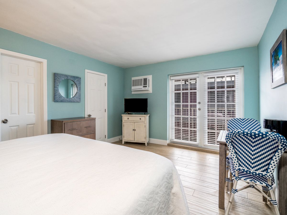 A bedroom with blue walls features a bed, dresser, TV, wall mirror, small desk with chairs, and a window with white shutters.