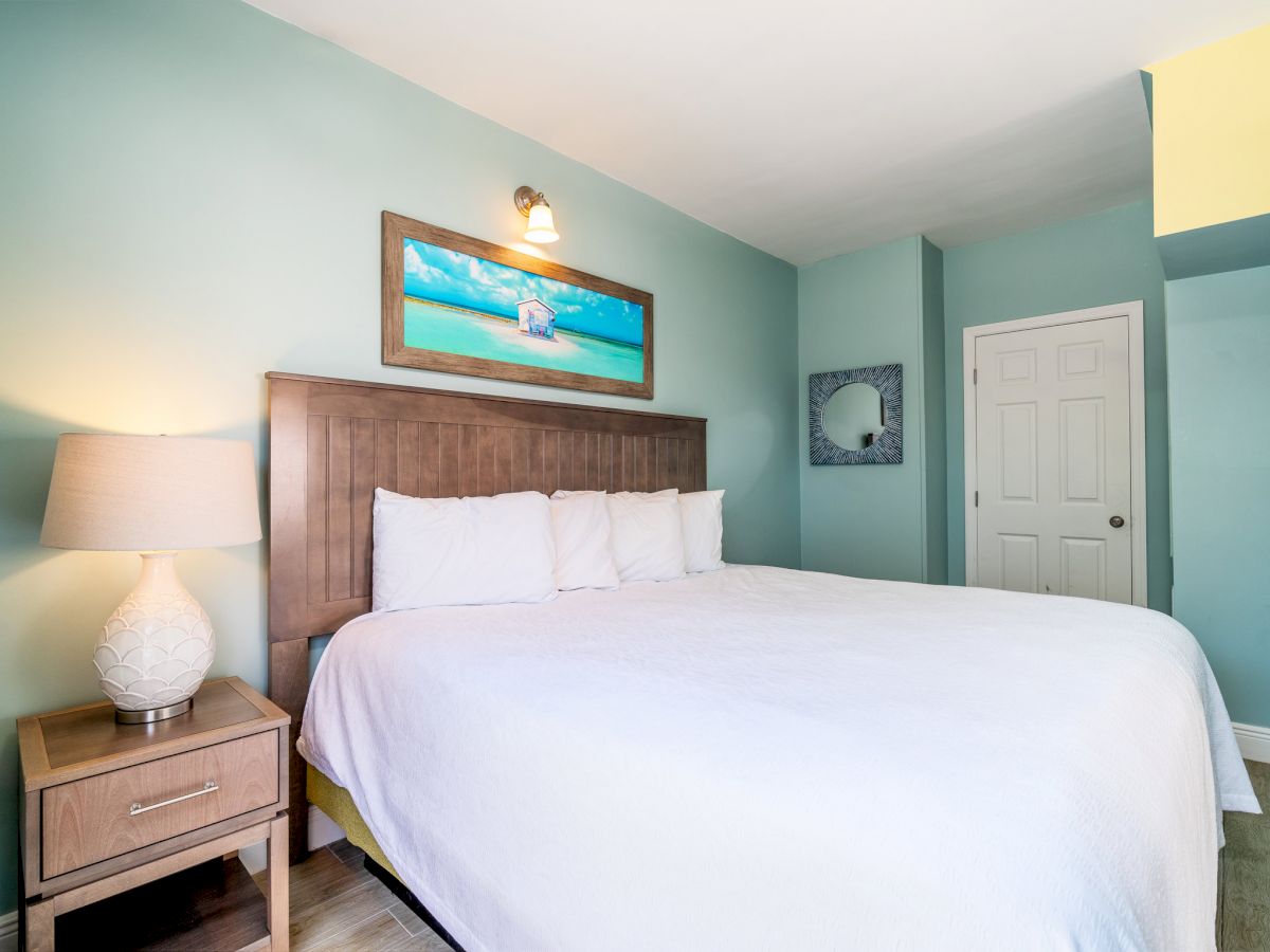 A bedroom with a large bed, light blue walls, a wooden nightstand with a lamp, a framed beach picture, and a door.