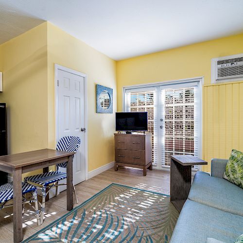 A small, colorful living area with a blue sofa, table, chairs, TV, and decorative elements, featuring bright yellow walls and natural light.