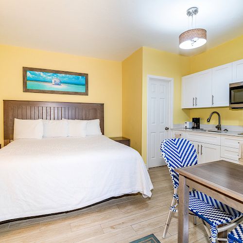 A cozy room with a bed, kitchenette, and dining area. Bright yellow walls, scenic artwork, and blue patterned chairs add charm.