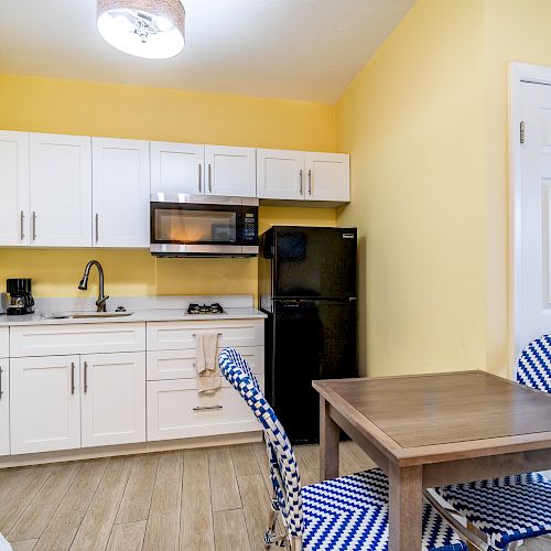 A small kitchen with white cabinets, a black fridge, microwave, and a wooden table with blue chairs, on a yellow wall background.