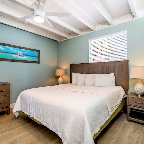 A cozy bedroom with a bed, two lamps, a dresser, and a beach-themed wall painting. The room has teal walls and wooden flooring.