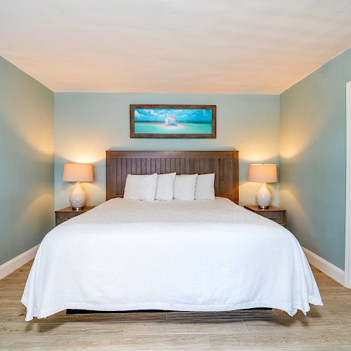 A bedroom with a bed, white bedding, two lamps on side tables, and a beach painting above the headboard.