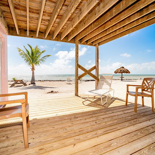 A wooden deck with chairs overlooks a sandy beach, palm trees, and the ocean under a clear blue sky.