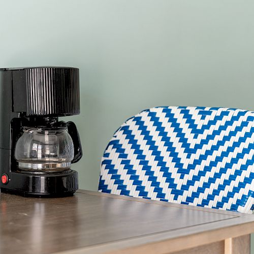 A black coffee maker sits on a wooden table next to a chair with a blue and white woven zigzag pattern.