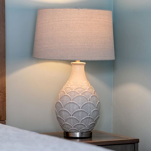 A decorative table lamp with a textured base and a gray shade on a bedside table in a room with light blue walls.