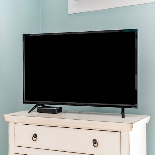 A flat-screen TV on a wooden dresser with a small device next to it, against a light blue wall.