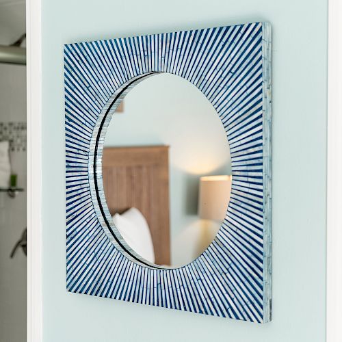 A round mirror with a blue, striped frame is on a wall. A reflection of a bed and lamp is visible in the mirror.