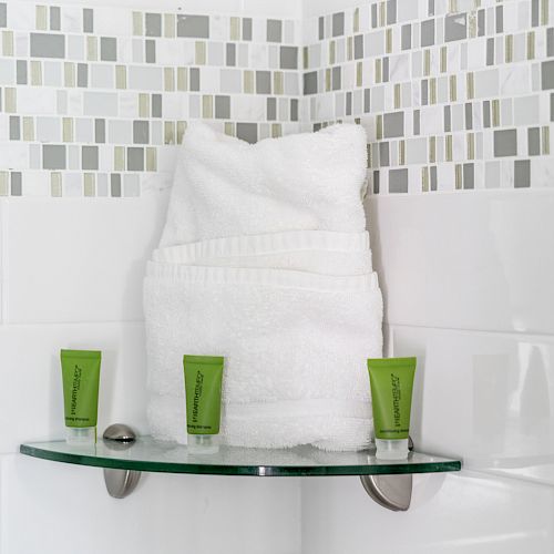 A glass shelf with folded white towels and three small green toiletry tubes in a bathroom with tiled walls.