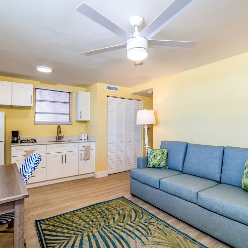 A cozy living room with a sofa, kitchen area, dining table, and ceiling fan; decorated with a tropical rug and leaf-patterned pillows.