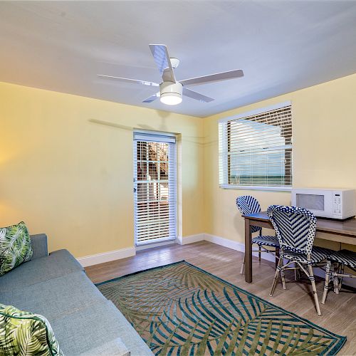 A cozy room with yellow walls, blue sofa, patterned rug, dining table with chairs, and a white microwave, under a ceiling fan.