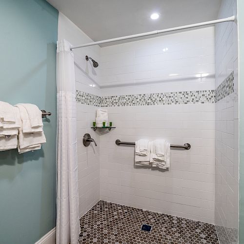 A bathroom with a white tiled shower, grab bar, towels, and a light blue wall. The floor has hexagonal tiles and there's a shower curtain.