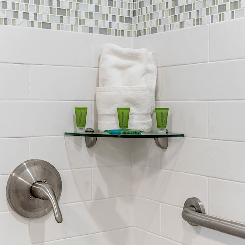 A bathroom corner with towels, small toiletry bottles, a soap on a glass shelf, and shower controls on white tiled walls.