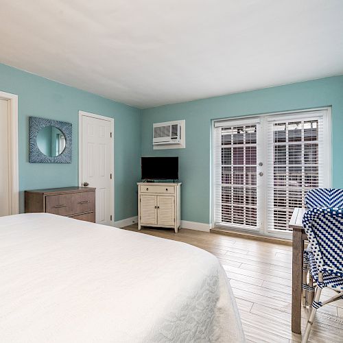 A bedroom with blue walls features a bed, wicker chairs, dresser, mirror, TV, air conditioner, and glass doors leading to outside.