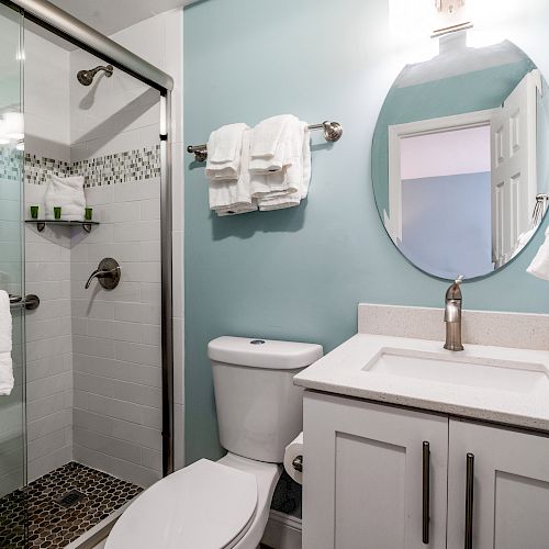 This image shows a modern bathroom with a shower, white towels, a circular mirror, and a sink with a light blue wall color.