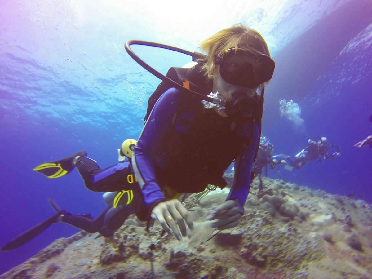 A scuba diver in blue gear is underwater, hovering above a coral reef, with other divers visible in the background.