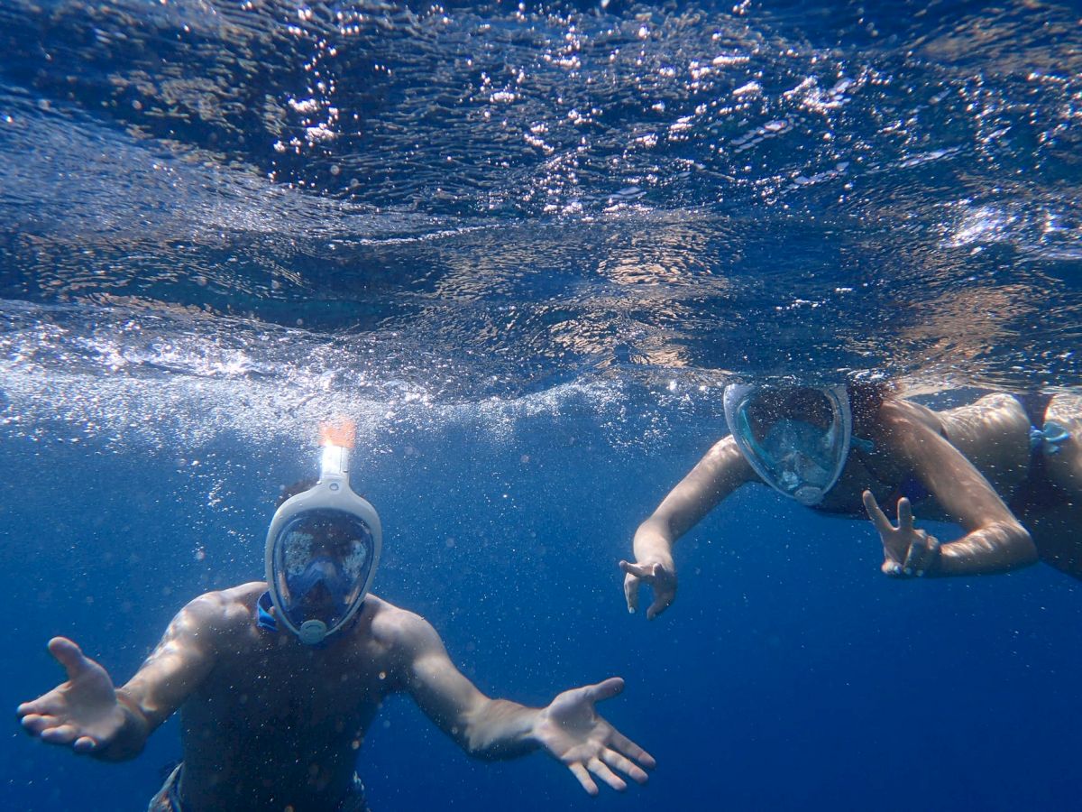 Two people are snorkeling underwater, wearing full-face masks, with one making a peace sign.