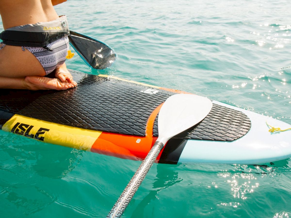 A person is kneeling on a paddleboard with a paddle resting on it, floating in clear blue water. The paddleboard has vibrant colors and the brand 