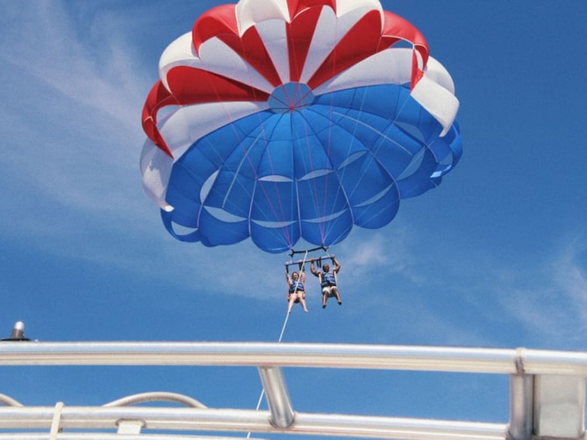 Two people parasailing with a red, white, and blue parachute are seen against a clear blue sky, with rooftops or boat structures in view below them.