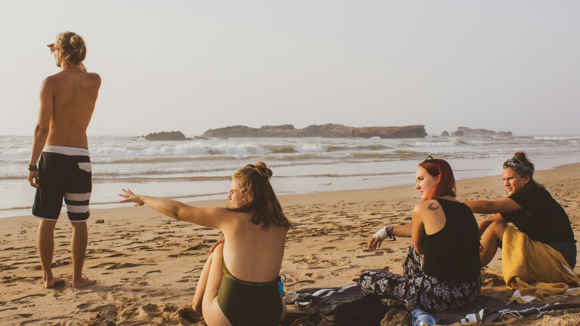 A group of four people relax on a sandy beach, with two seated women, a standing man, and another gesturing woman facing the ocean waves.