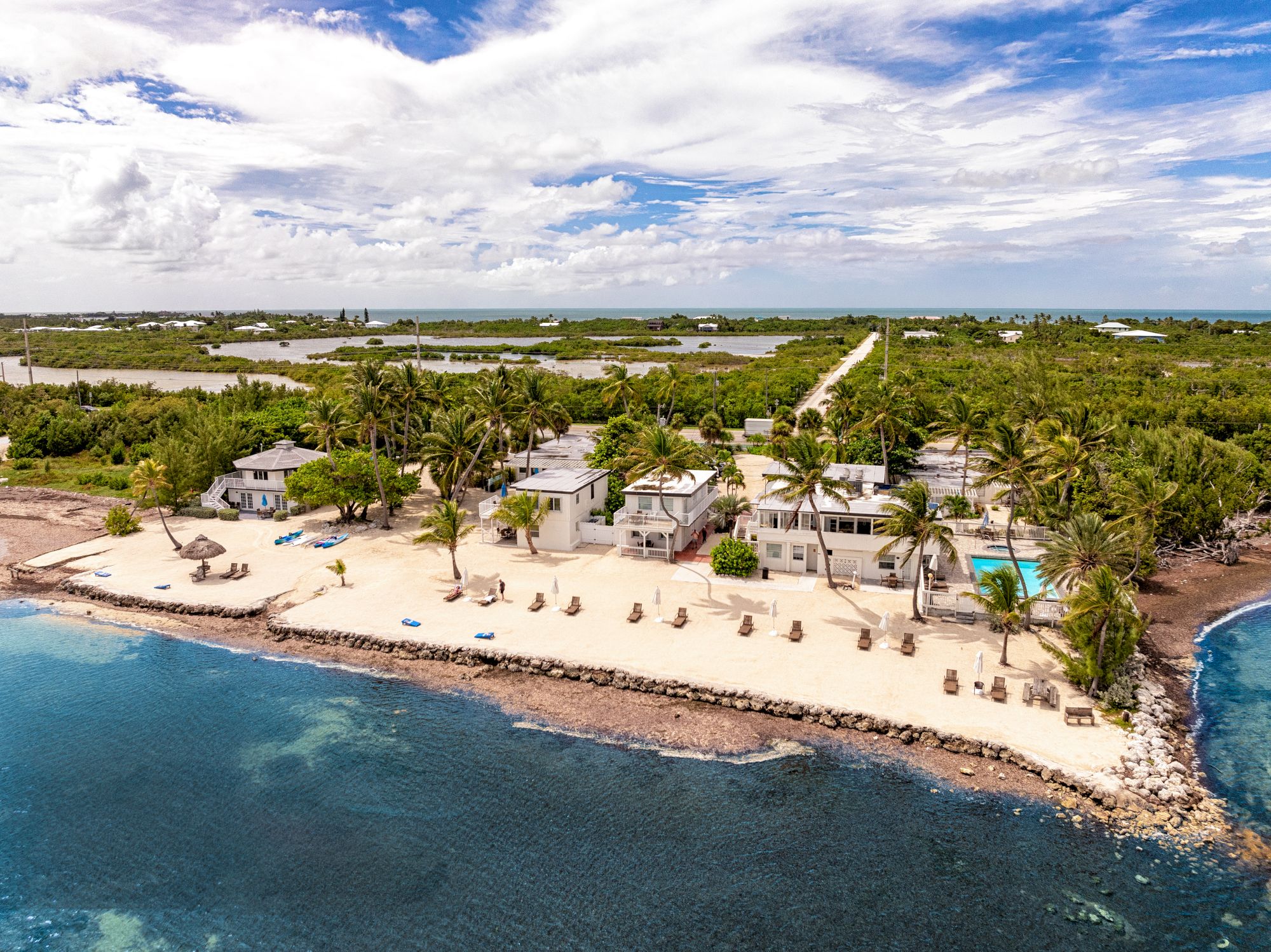 A beachfront property is surrounded by palm trees, sandy shores, and a pool, with a road leading into lush greenery and coastal waters.