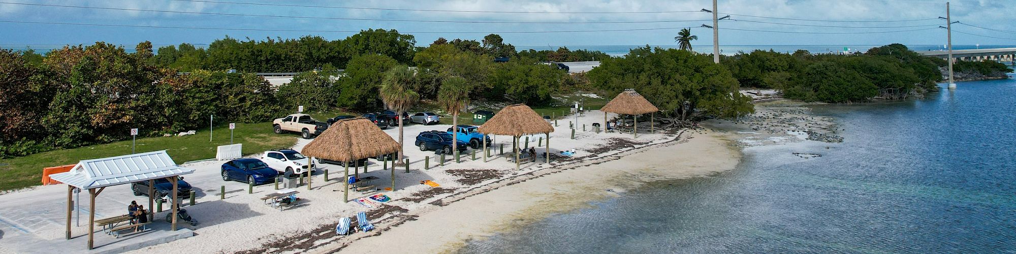 A serene beach with huts and parked vehicles, bordered by lush greenery and calm water under a sky dotted with fluffy clouds.