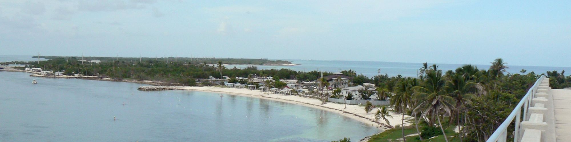 A coastal view from a bridge, showing palm trees, a curved beach, and a distant shoreline under a cloudy blue sky.