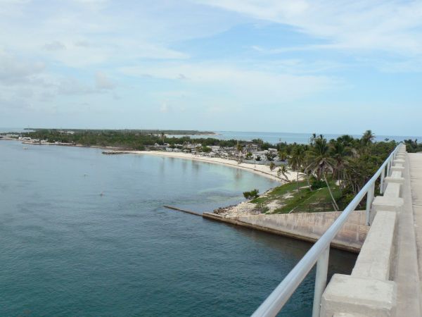 View from a bridge overlooking a coastline with clear blue water, a sandy beach, and palm trees under a partly cloudy sky.