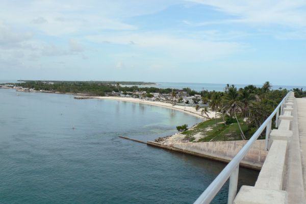 A coastal landscape view from a bridge, overlooking the ocean and a beach with trees and buildings in the distance.
