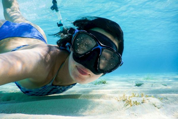 A person is snorkeling underwater, wearing a blue swimsuit and snorkel mask, swimming above sandy ocean floor with clear blue water.